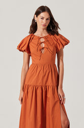 A woman is wearing a coral-colored dress with puffed short sleeves, a fitted bodice with a lace-up front featuring multiple bows, and a tiered skirt with a slit on one side.